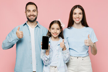 Wall Mural - Young fun parents mom dad with child kid daughter teen girl in blue clothes hold use mobile cell phone screen workspace area show thumb up isolated on plain pastel pink background. Family day concept.