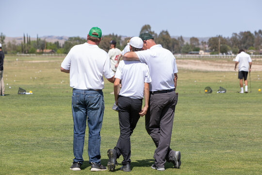Looking at the back of three men standing on the golf course. One man talking to another with his arm around his shoulder. Golf coach or father giving instruction.