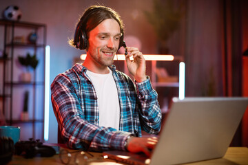 Wall Mural - Portrait of young caucasian man in headset sitting at desk and using modern laptop for remote work at home. Male freelancer looking at laptop screen with serious facial expression.