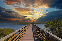 A Long Brown Wooden Bridge Over A Lush Green Marsh Surrounded By Vast Blue Ocean Water With Powerful Clouds At Sunset At Bolsa Chica Ecological Reserve In Huntington Beach California USA