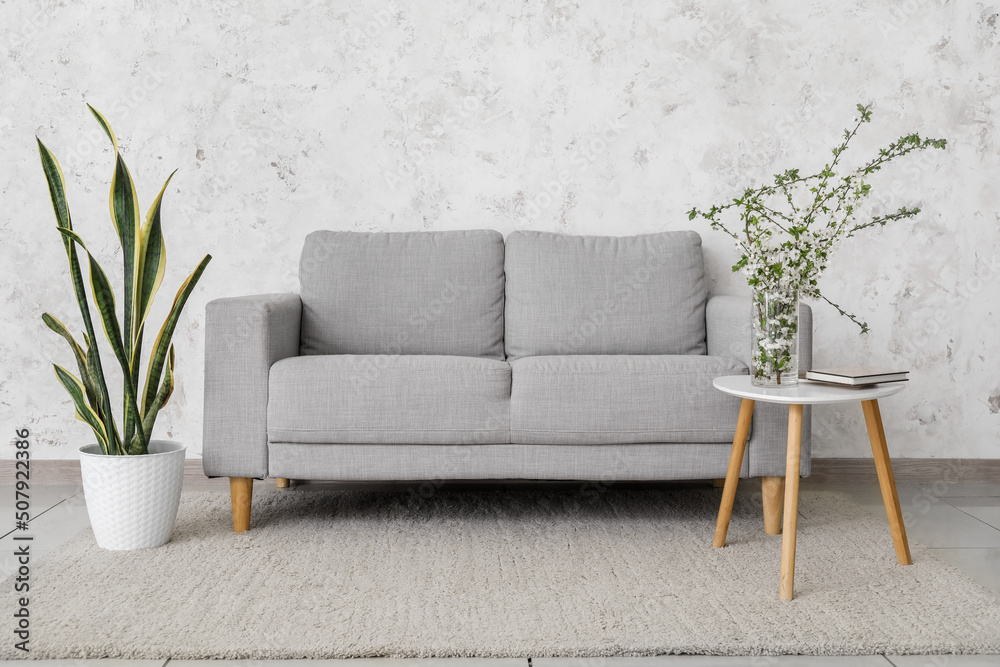 Vase with blooming branches, grey sofa and houseplant near light wall