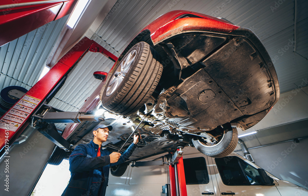 Young car mechanic at repair service station inspecting car wheel and suspension detail of lifted automobile. Bottom view.