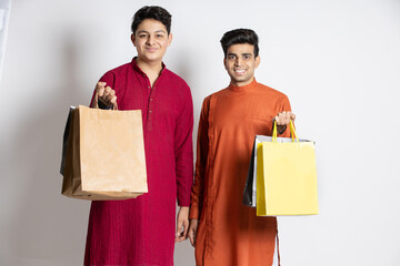 Wall Mural - Two Happy young indian men holding shopping bags. friends or brother shopper wearing ethnic outfit,isolated over white studio background, black Friday deal, diwali festive sale discount.