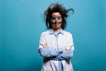 Hilarious Looking Crazy Chemist With Messy Hairstyle And Dirty Face Standing On Blue Background Looking At Camera. Humorous Mad Scientist With Erratic Hair And Filthy Face Standing With Crossed Arms.