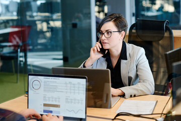 Young businesswoman in eyeglasses having a conversation on mobile phone while working at her workplace with laptop