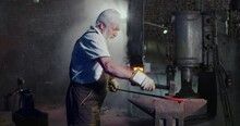 Side View Of Skillful Blacksmith Working In Mastershop. Old Strong Male With Grey Hair And Beard Red Stick By Hands Holding, With Hammer Beating. Concept Of Blacksmith Art.