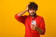 Thoughtful bearded man scratching head holding smartphone in hand, contemplating about software updating, choosing suitable tariffs. Indoor studio shot isolated on orange background