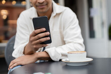 Cropped View Of Blurred African American Man Texting On Smartphone Near Cup Of Cappuccino On Table On Cafe Terrace.