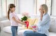 Cute little daughter is congratulating mom and giving her flowers roses and handmade postcard. Attentive small kid made gift on International Women Day to loving mom, Mother Day celebration.