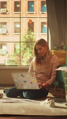 Poster - Vertical View of Blonde Woman Using Laptop Computer, while Sitting on Living Room Floor in Cozy Stylish Apartment. Attractive Girl is Pondering and Coding