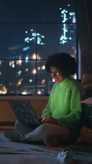 Poster - Positive Latina Female Using Laptop with Stickers at Home. Woman Resting at the Floor and Messaging at Computer with Smile. Evening View Behind the Window. Vertical Shot