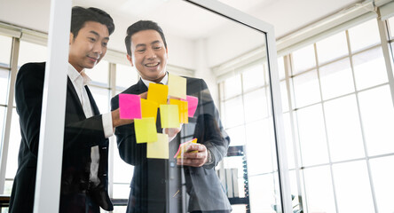 Group of successful Asian businessmen teamwork. Brainstorm meeting with sticky paper notes on the glass wall for new ideas. Using agile methodology for business in a tech start-up office.