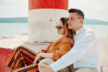 A Couple Sits Near A Lighthouse On The Beach Against The Backdrop Of The Sea And Mountains. Vacation Love, Honeymoon, Travel. Stylish Couple.