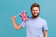 © khosrork - Portrait of handsome satisfied bearded man holding flag of a constituent unit of the United Kingdom, celebrating British Independence Day. Indoor studio shot isolated on blue background.