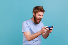 Portrait Of Handsome Young Adult Bearded Man Standing, Using Smartphone And Playing Mobile Game With Excited Positive Face, Happy To Win. Indoor Studio Shot Isolated On Blue Background.