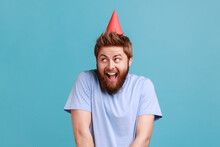 Portrait Of Pleasant Surprised Handsome Bearded Man Standing Happy Facial Expression, Celebrating Her Birthday With Party Cone On His Head. Indoor Studio Shot Isolated On Blue Background.