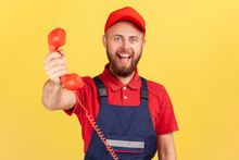 Portrait Of Positive Handyman Wearing Blue Uniform Holding Out Handset, Asking Call Him And Order Service, Looking At Camera Happy Expression. Indoor Studio Shot Isolated On Yellow Background.