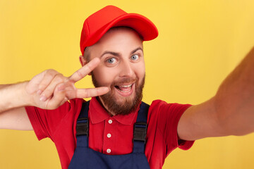 Wall Mural - Smiling positive man courier or repairman broadcasting livestream or making point of view photo, showing v sign, wearing blue overalls and red cap. Indoor studio shot isolated on yellow background.