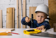 Little Boy With Helmet Learn To Hammer A Nail In Wooden Plank At Carpenters Workshop
