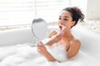 Beautiful young lady shaving off unwanted hair on face while taking bubble bath at home, free space