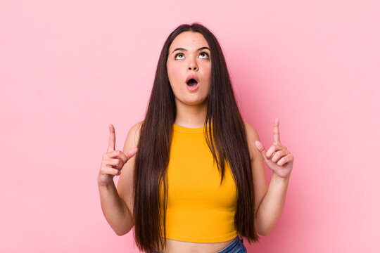 young adult woman looking shocked, amazed and open mouthed, pointing upwards with both hands to copy space
