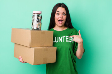 Wall Mural - young adult woman looking shocked and surprised with mouth wide open, pointing to self. donation concept