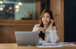 © crizzystudio - Happy business asian manageress working at her desk in the office taking a call on her mobile phone while writing notes on a notepad