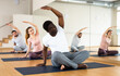 Adult African American practicing yoga during group class in fitness studio, doing side bend stretching sitting in Padmasana pose..