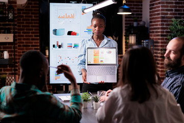 African american woman standing in business office presenting business analytics on laptop screen in late night meeting. Startup entrepreneur discussing sales charts with coworkers working overtime.