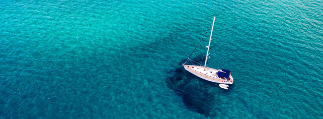 Aerial view of anchored sailing yacht in emerald sea. Aerial view of a boat. Outdoor water sports, yachting. Aerial view of anchoring yacht in open water. Ocean and sea travel and transportation