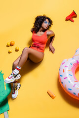 Wall Mural - high angle view of african american woman in swimsuit near swim ring, fruits and loudspeaker on yellow background.