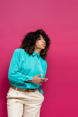 Wall Mural - african american woman in turquoise blouse standing with hand in pocket and looking away isolated on pink.