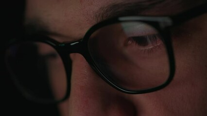 Close up of focused male hacker wearing computer glasses for reducing eye strain. Young man looking at screen with computer reflection in dark room. Concept of cyber crime.