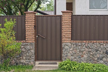 Poster - one brown metal door on the wall of a fence made of bricks and iron on a gray stone foundation in a street in green vegetation