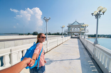 Wall Mural - Follow me. Travel concept. Vacation on Phuket island. Back view of young woman holding boyfriend's hand enjoying sea view from bridge terrace.