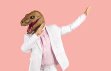Funny Weird Guy In Wacky Animal Mask Having Fun At Crazy Party. Eccentric Man In White Suit And Silly Ugly Masquerade Dinosaur Mask Dancing Isolated On Pink Background