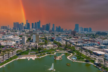 an aerial shot of silky green lake in the park surrounded by lush green palm trees and grass with skyscrapers and office buildings in the city skyline with powerful clouds at sunset and a rainbow