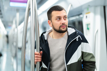 Portrait of thoughtful young bearded man holding on handrails in subway car during daily commute to work ..