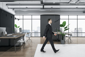 Poster - Attractive thoughtful young european businessperson walking in wooden and concrete office interior with furnituire and window with city view. CEO, executive and consulting concept.