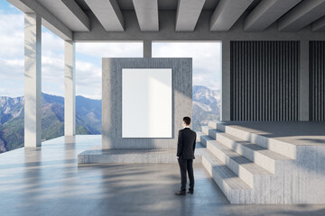 Poster - European businessman standing in bright modern concrete terrace or balcony interior with empty white mock up poster, beautiful nature view and bright sky. Architecture and design concept.