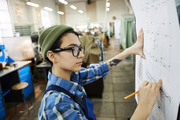 Wall Mural - Serious busy tenacious lady in glasses standing at wall and drawing on blueprint while working with technical card