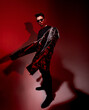 Front view of brutal male model in sunglasses holding stylish jacket in hands, standing in isolated studio with red light, showing collection of designer apparel