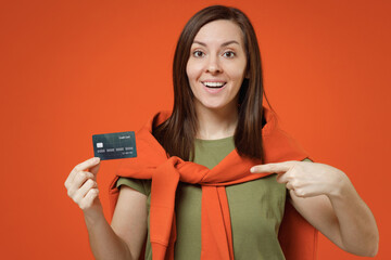 Wall Mural - Young smiling fun happy woman 20s in khaki t-shirt tied sweater on shoulders hold point index finger on credit bank card isolated on plain orange background studio portrait. People lifestyle concept.