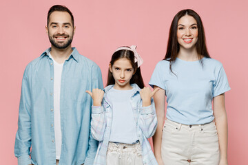 Wall Mural - Young happy smiling fun cool parents mom dad with child kid daughter teen girl in blue clothes point thumb fingers on mother fater isolated on plain pastel light pink background. Family day concept.