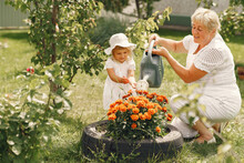 Little Girl And Her Grandmother Watering Flowers In Garden