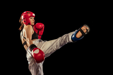 Wall Mural - High kick. Attractive female boxer in boxing gloves and helmet training isolated on dark studio background. Sport, competition, hobby, results, success concept