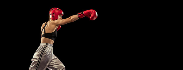 Wall Mural - Young female boxer in boxing gloves and helmet training isolated on dark studio background. Sport, achievements, motivation, hobby, results, success concept