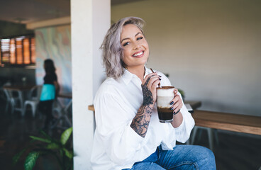 Portrait of funny female holding chocolate frappe beverage and smiling at camera during weekend leisure spending in local cafe interior, cheerful Caucaisan hipster girl with non caffeine latte