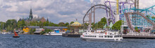 Small Open Sailing Boat With Red Sail, A Commuter Ferry And Commuter Boat In A Bay At An Amusement Park A Sunny Day In Stockholm