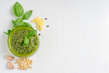 Wall Mural - Top view of italian herbal homemade pesto sauce made of blended green basil leaves, parmesan cheese, pine nuts, garlic and olive oil served in glass bowl on white wooden background with copy space